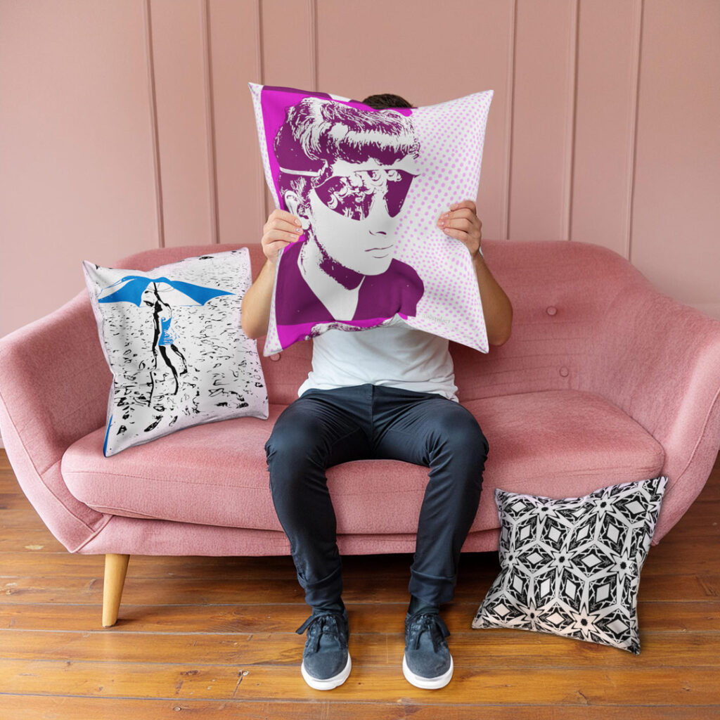 A young man sitting on a couch holds a pillow in front of his face. The pillow has another face on it, as if to look like the pillow is the face of the man. There are other pillows around the man on the counch.